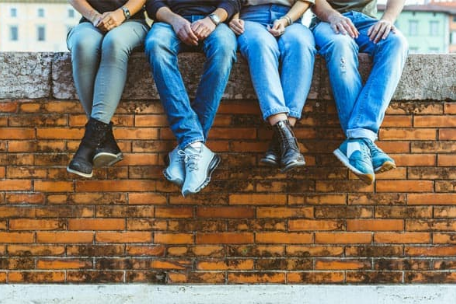 Four pairs of legs in jeans with different shoes sitting on a brick wall.