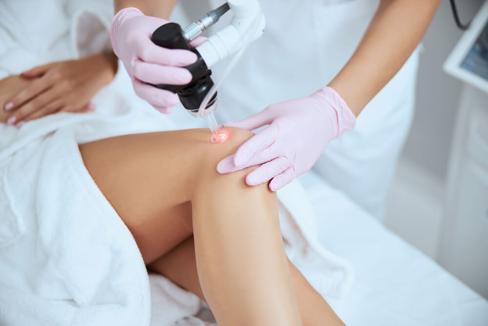 Patient in white robe receiving professional laser vein treatment for varicose veins on knee from specialist wearing pink gloves at Nelson Vein Clinic in Cleveland, Ohio, modern medical clinic setting.